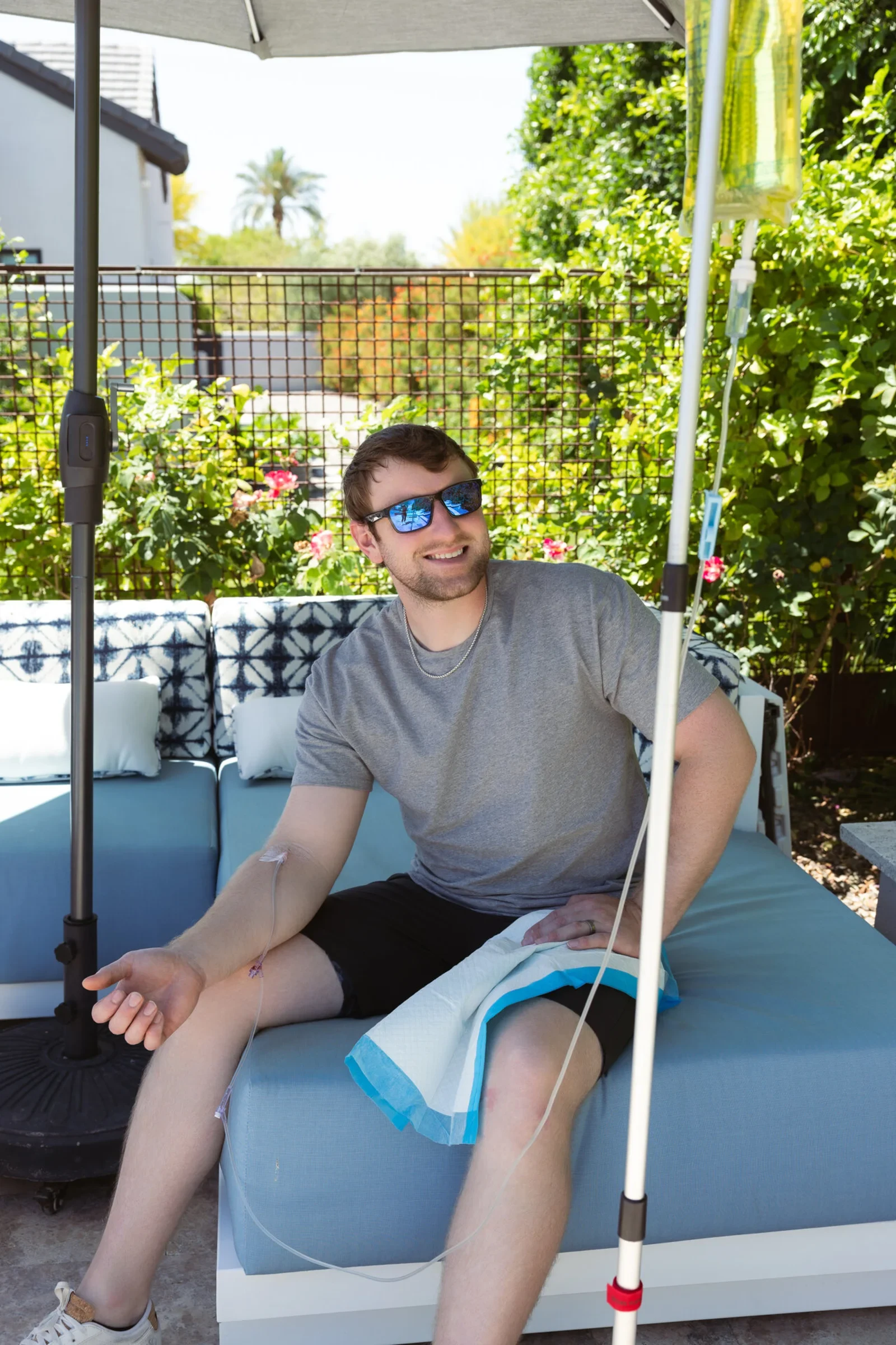 A man in sunglasses on a shaded patio daybed in Phoenix, receiving an IV with the stand beside him