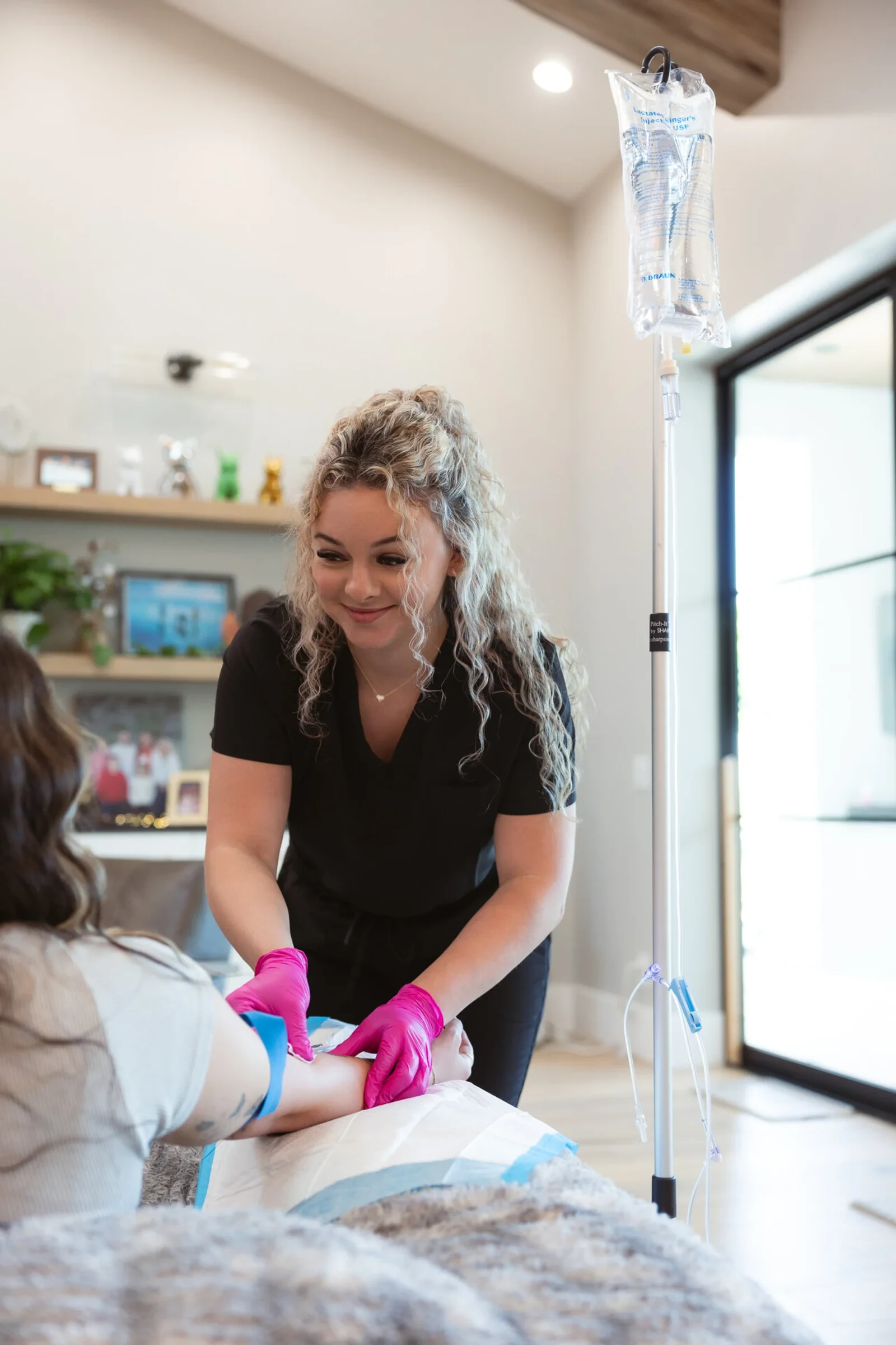 A registered nurse in black scrubs and pink gloves starting a client's IV in a bright Phoenix living room, the bag already hanging on its stand