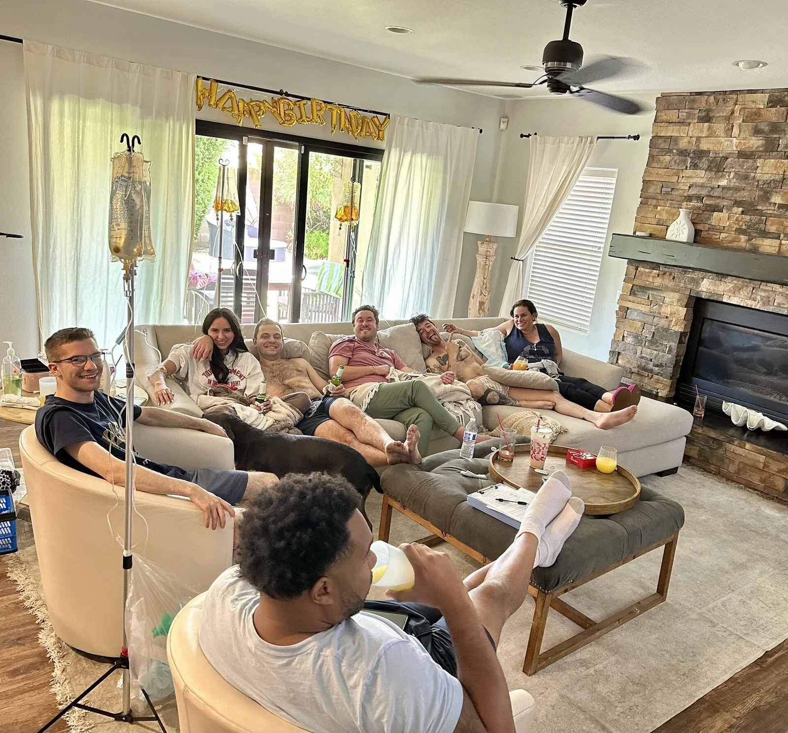A birthday group at a Phoenix home, five friends spread across the sofas on drips with stands beside them and a banner over the window