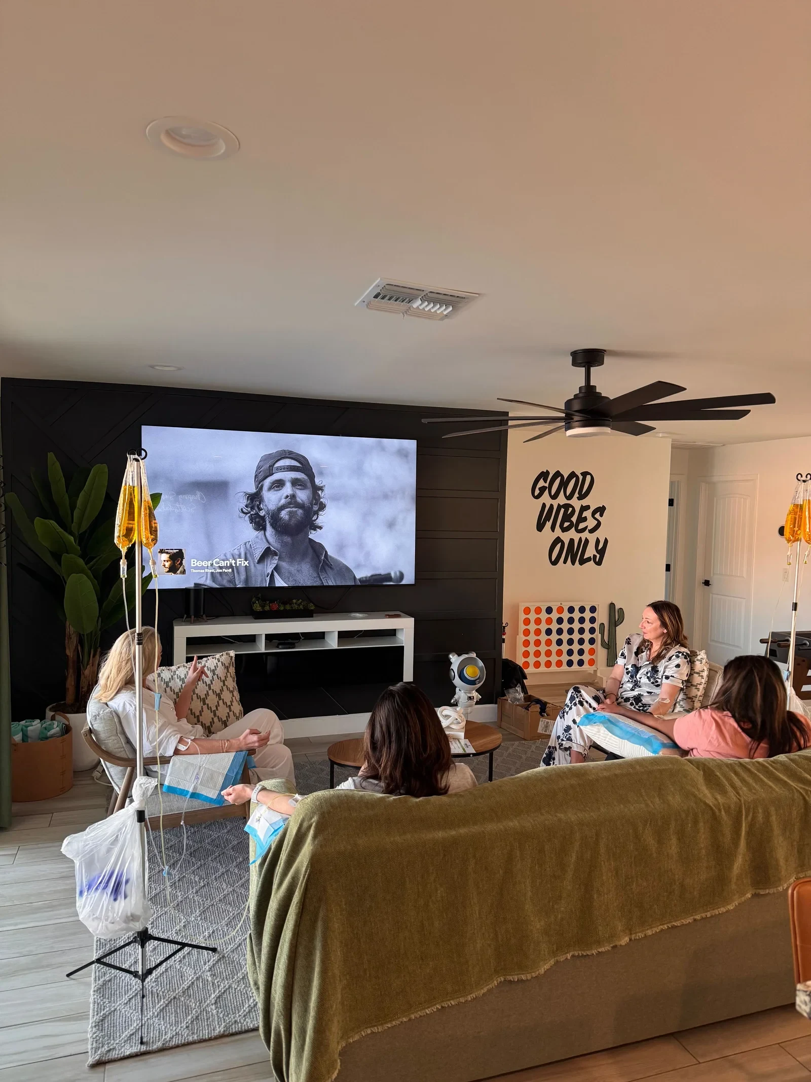 A group of friends on drips in a Phoenix living room, IV stands lined up beside the sofa and a film on the television