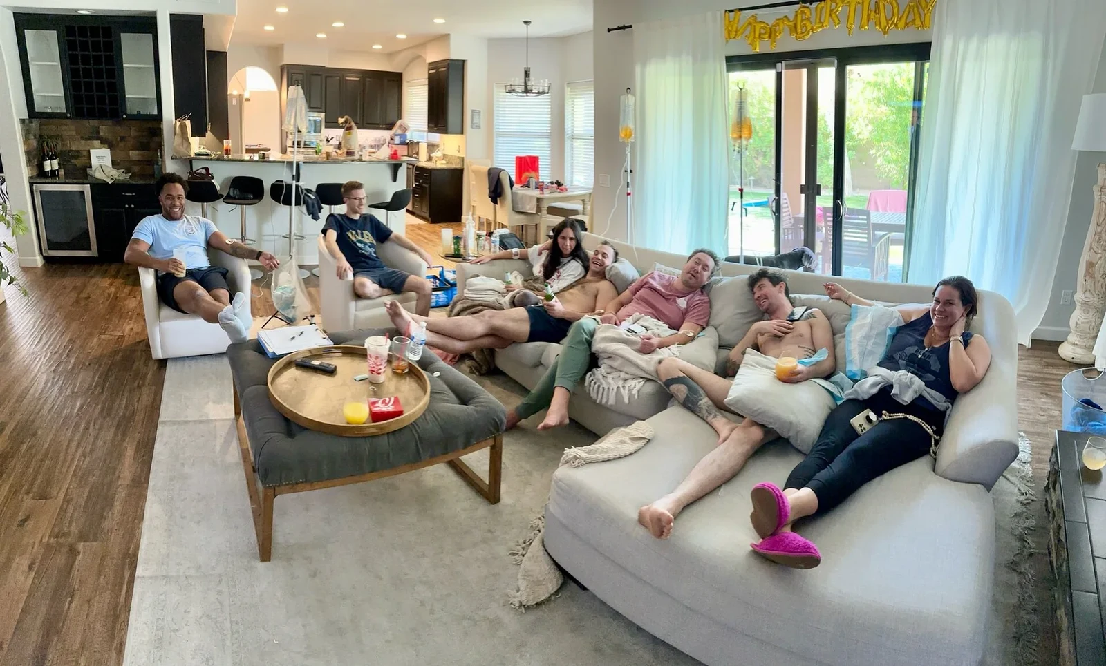 A full living room of friends on drips at a Phoenix house, stands between the sofas and a banner across the window
