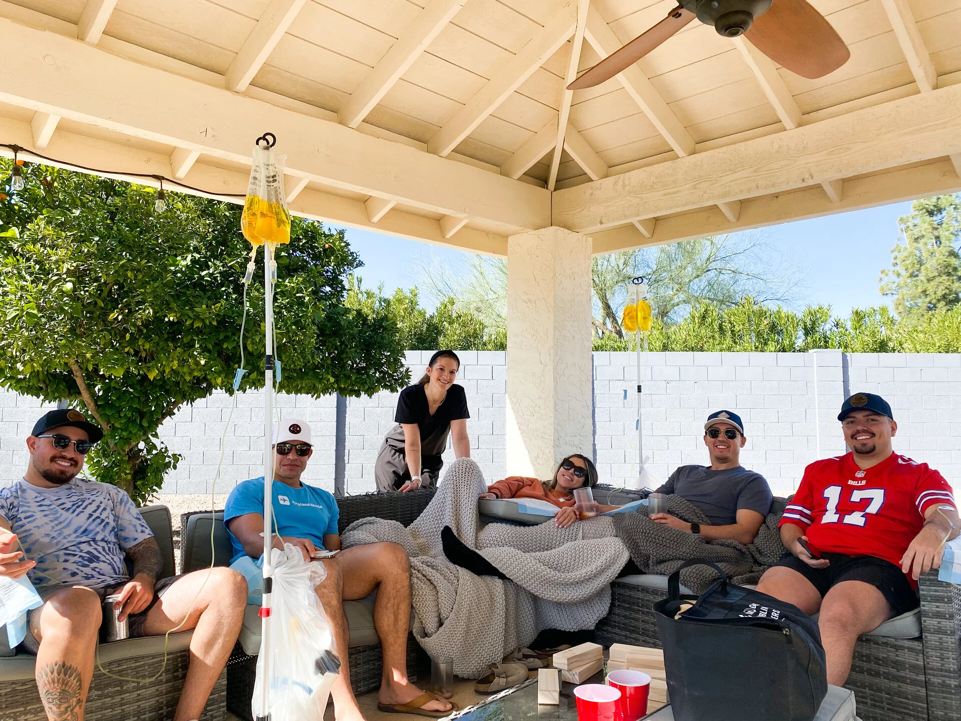 Friends on a shaded Phoenix patio under a wooden pergola, each on a drip, grinning at the camera with blue sky and desert trees behind