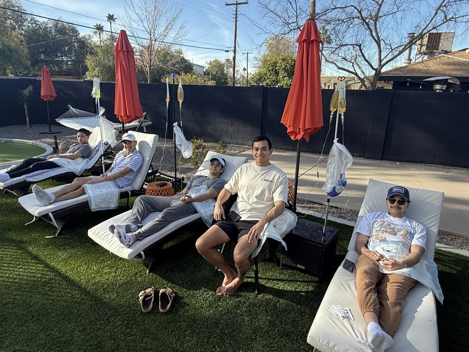 Five friends on loungers beside a Phoenix pool, each on a drip with a stand behind them and parasols overhead