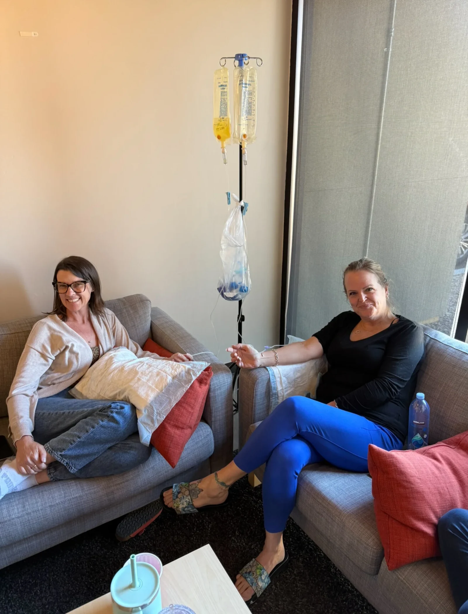 Two women side by side on the sofa at home, both on drips from the same stand between them