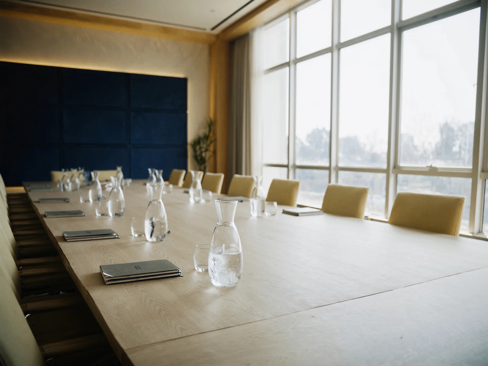 A sunlit boardroom set for an offsite: a pale oak table, carafes of iced water and closed notebooks by tall windows