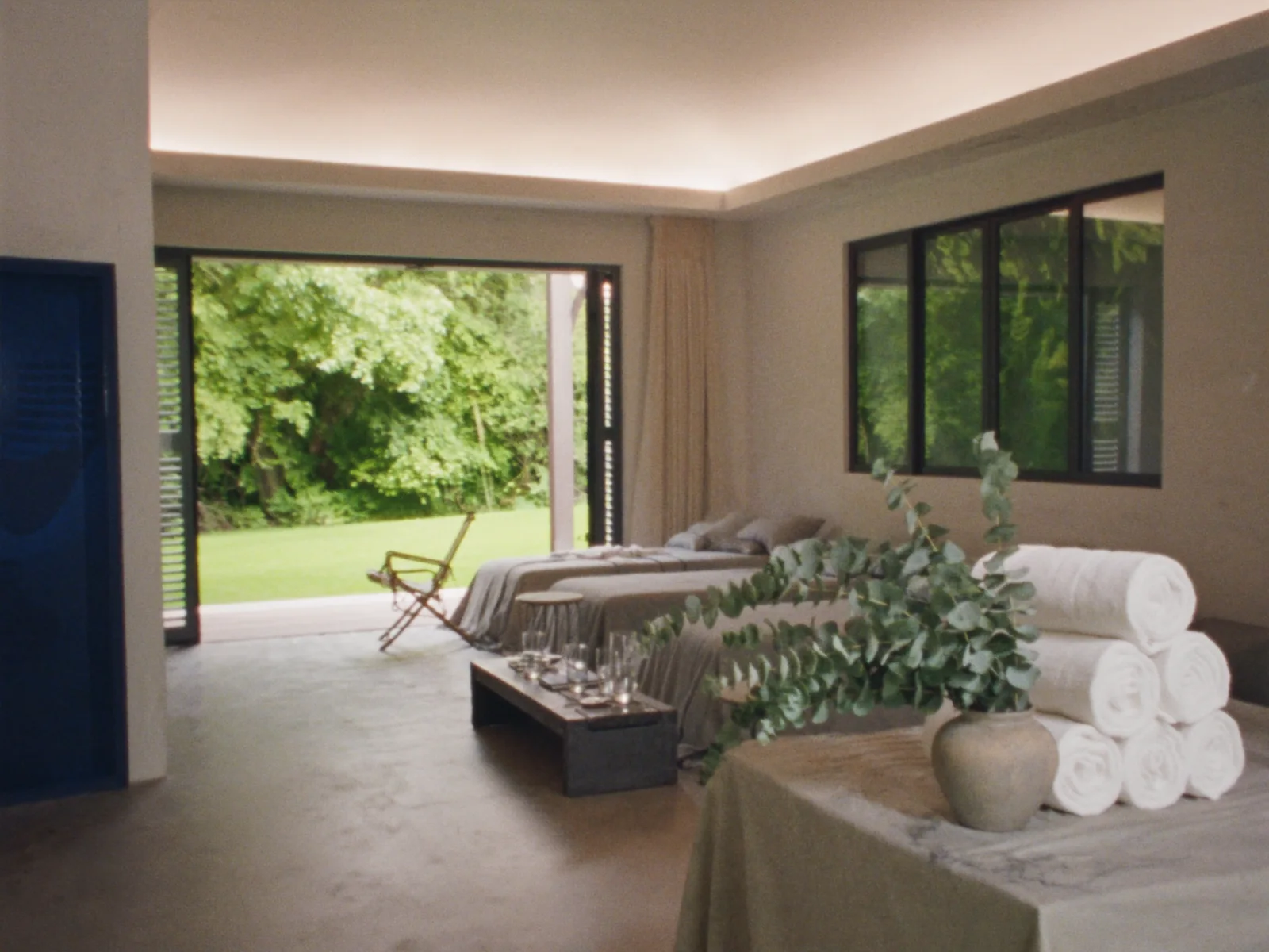 A retreat lounge with pale linen daybeds, folded towels, a glass pitcher and eucalyptus, opening onto greenery
