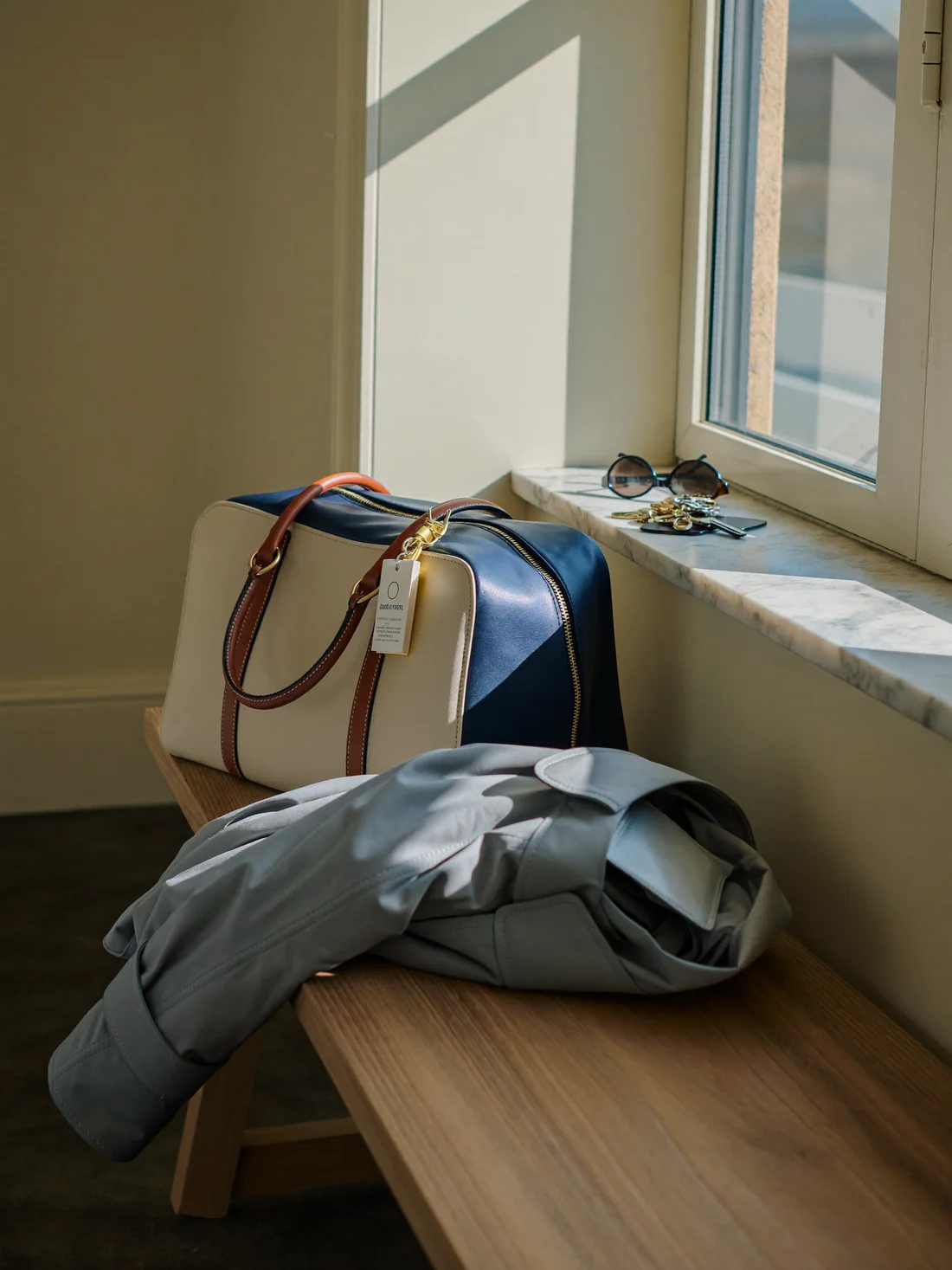 A leather weekender bag and folded coat on a bench in a sunlit entry hall