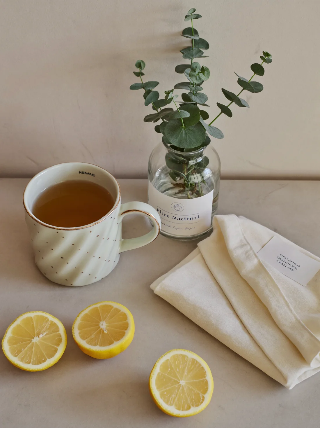 A steaming mug of tea with halved lemons and eucalyptus on a pale marble counter