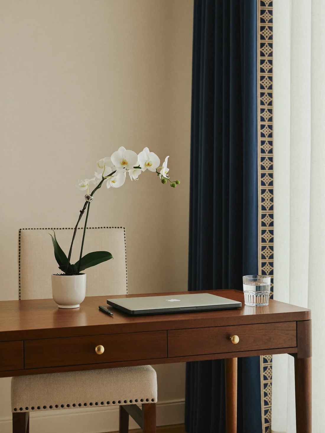 A calm home desk with a closed laptop, a glass of water and a white orchid by a window