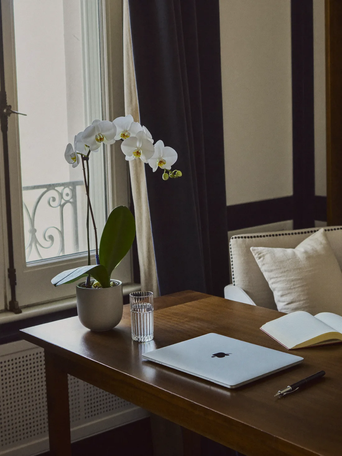A calm home desk with a closed laptop, a glass of water and a white orchid by a window